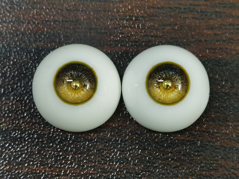 Puede incluir: Dos ojos de mu&ntilde;eca realistas con escler&oacute;tica blanca, iris negro y pupila dorada y marr&oacute;n. Los ojos est&aacute;n colocados sobre un fondo marr&oacute;n.