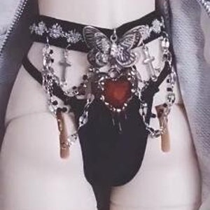 K&ouml;nnte beinhalten: Schwarze Dessous mit einer silbernen Kette und Kreuz-Akzenten, einem gro&szlig;en silbernen Schmetterlingsanh&auml;nger und einem roten Herz-Anh&auml;nger.
