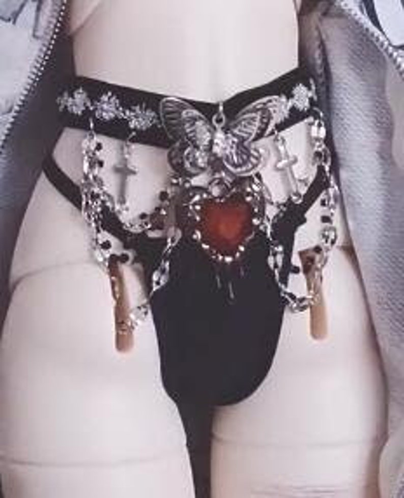 K&ouml;nnte beinhalten: Schwarze Dessous mit einer silbernen Kette und Kreuz-Akzenten, einem gro&szlig;en silbernen Schmetterlingsanh&auml;nger und einem roten Herz-Anh&auml;nger.