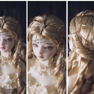 May include: A doll with long blonde hair wearing a gold-toned headpiece featuring a central blue stone, dangling chains, and small star and moon charms. The doll also wears a matching gold-toned necklace adorned with white pearls and small blue stones.
