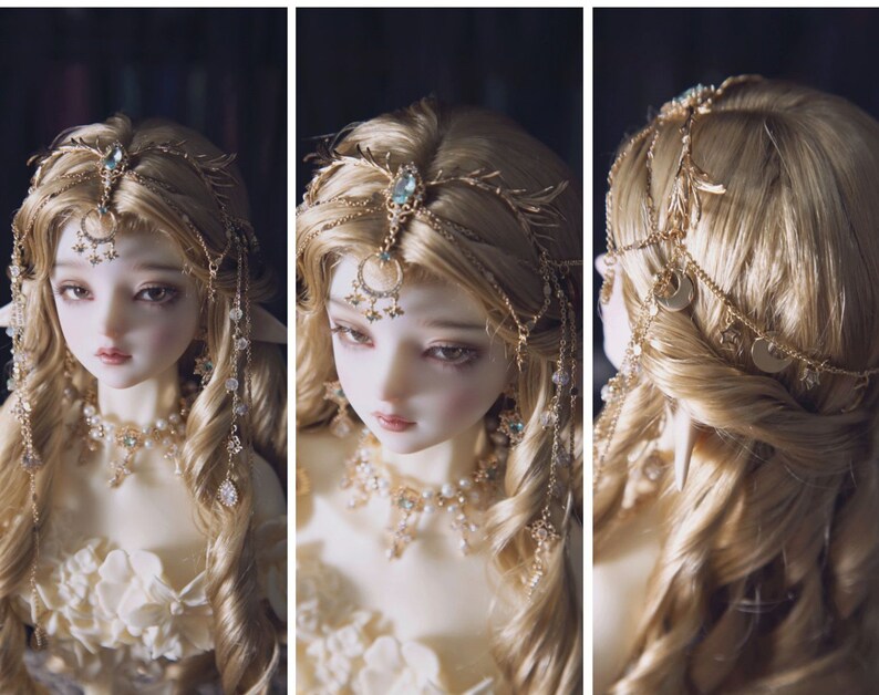 May include: A doll with long blonde hair wearing a gold-toned headpiece featuring a central blue stone, dangling chains, and small star and moon charms. The doll also wears a matching gold-toned necklace adorned with white pearls and small blue stones.