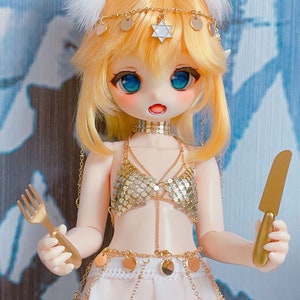 May include: A doll with blonde hair and blue eyes is wearing a gold chainmail top, a white skirt, and a gold belt with coin accents. The doll is holding a fork in one hand and a knife in the other.