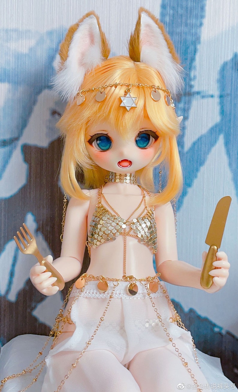 May include: A doll with blonde hair and blue eyes is wearing a gold chainmail top, a white skirt, and a gold belt with coin accents. The doll is holding a fork in one hand and a knife in the other.