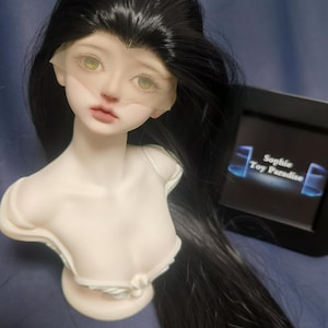 May include: A porcelain doll head with long black hair and a white lace veil. The doll has green eyes and is wearing a white dress. The doll is sitting on a blue surface. The text "Sophie Toy Paradise" is visible in the background.