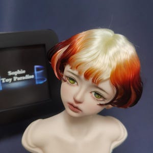 May include: A doll head with a short, layered wig. The wig is a blend of blonde, orange, and red hair. The doll has green eyes and a neutral expression.