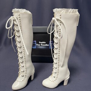 Pode incluir: Um par de botas brancas, at&eacute; ao joelho, com detalhes de atacadores e saltos pequenos. As botas t&ecirc;m uma guarni&ccedil;&atilde;o decorativa na parte superior e s&atilde;o feitas de um material liso. As botas s&atilde;o exibidas numa superf&iacute;cie escura.