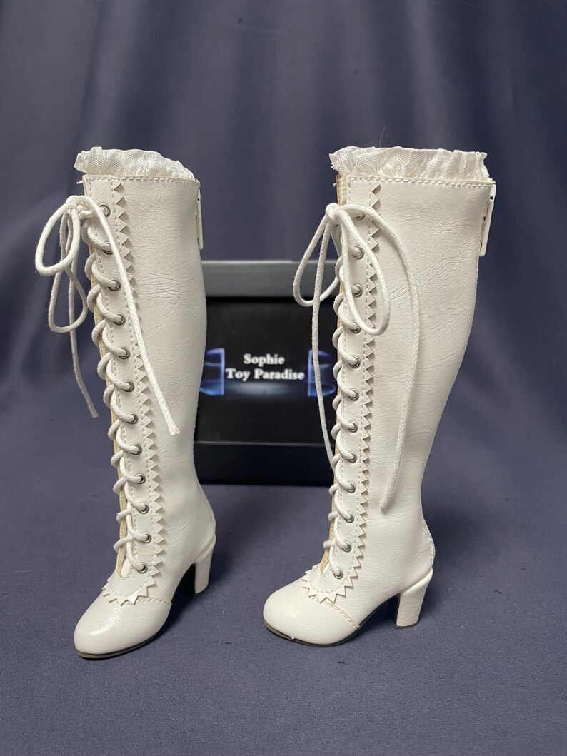 Pode incluir: Um par de botas brancas, at&eacute; ao joelho, com detalhes de atacadores e saltos pequenos. As botas t&ecirc;m uma guarni&ccedil;&atilde;o decorativa na parte superior e s&atilde;o feitas de um material liso. As botas s&atilde;o exibidas numa superf&iacute;cie escura.