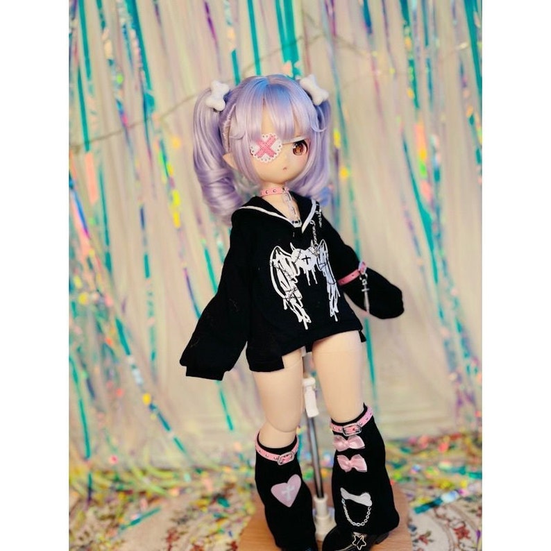 K&ouml;nnte beinhalten: Eine Puppe mit lila Haaren, die ein schwarzes Sweatshirt mit einem wei&szlig;en Skelettfl&uuml;gel-Design, ein rosafarbenes Augenpflaster und schwarze und rosafarbene Beinstulpen mit rosafarbenen Schleifen tr&auml;gt.