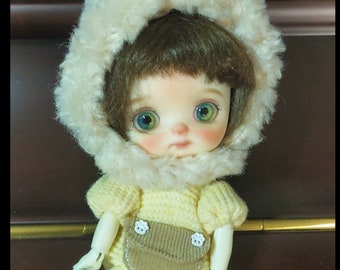 May include: A small doll wearing a yellow knitted outfit with a brown corduroy pocket and a white furry hat with bear ears. The doll has green eyes and is wearing yellow socks.