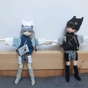 May include: Two dolls wearing different outfits. The doll on the left is wearing a white and blue sweater, black and white checkered shorts, and a white hat with ears. The doll on the right is wearing a black and white checkered shirt, black and white checkered shorts, and a black hat with ears.