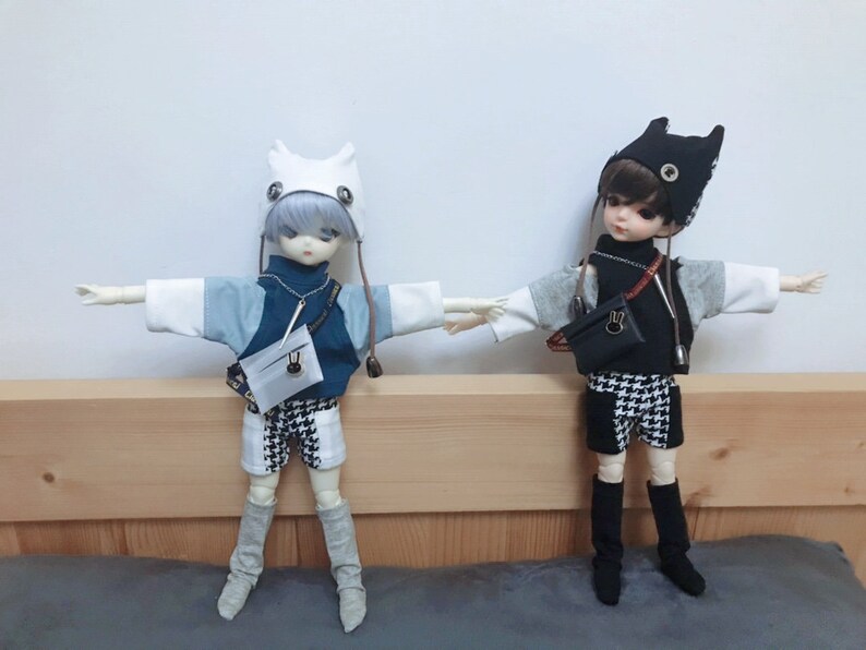 May include: Two dolls wearing different outfits. The doll on the left is wearing a white and blue sweater, black and white checkered shorts, and a white hat with ears. The doll on the right is wearing a black and white checkered shirt, black and white checkered shorts, and a black hat with ears.