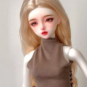 May include: A ball-jointed doll with pale skin, long blonde hair, and large eyes. The doll wears a sleeveless brown high-neck crop top featuring side lacing with metal eyelets. The doll has a neutral expression with defined lips and painted features.