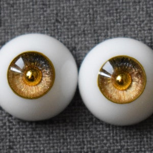 Puede incluir: Dos ojos de mu&ntilde;eca marrones y dorados con escler&oacute;tica blanca e iris dorado. Los ojos tienen un acabado brillante.