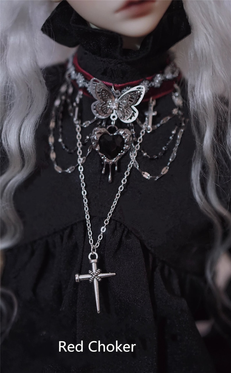 K&ouml;nnte beinhalten: Eine silberne Halskette mit einem schwarzen Herz-Anh&auml;nger und einem Kreuz-Anh&auml;nger. Die Halskette ist mit anderen silbernen Ketten und einem roten Choker mit einem silbernen Schmetterlings-Anh&auml;nger geschichtet.