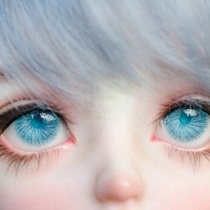 K&ouml;nnte beinhalten: Nahaufnahme eines Puppenkopfes mit blauen Augen und hellblauem Haar. Die Augen der Puppe sind offen und schauen direkt in die Kamera.