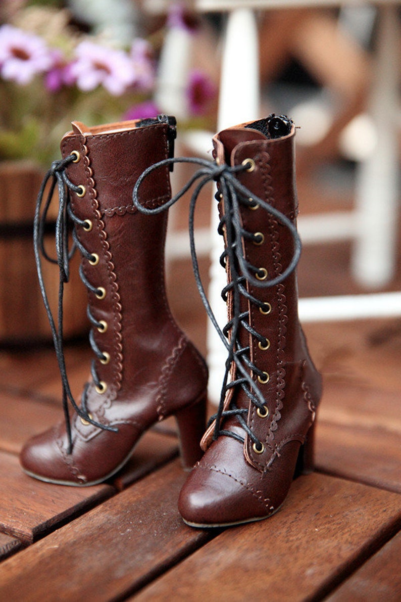 May include: A pair of brown leather boots with laces and a high heel. The boots have a scalloped edge design along the sides and gold-coloured eyelets. The laces are black, and the boots are on a wooden surface.