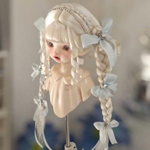 May include: A doll head with pale skin, blonde braided hair, and blue bows. The hair is styled with a jeweled headband and adorned with light blue ribbons and butterfly accents. The doll has delicate facial features and is mounted on a stand.