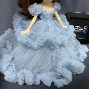 May include: A light blue tulle dress with a ruffled skirt and beaded bodice. The dress is designed for a doll and has a delicate, feminine look.
