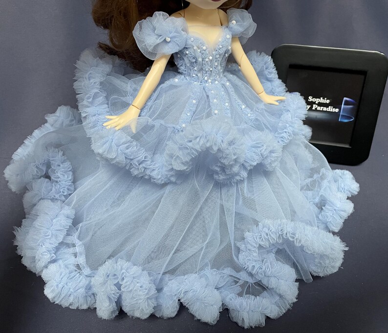 May include: A light blue tulle dress with a ruffled skirt and beaded bodice. The dress is designed for a doll and has a delicate, feminine look.