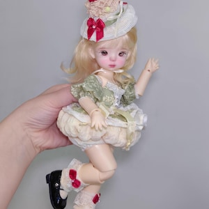 May include: A doll with blonde hair and a detailed face, wearing a green and cream-colored dress, black shoes, and a hat with a cherry on top. The doll is posed in a playful stance, with one arm raised.