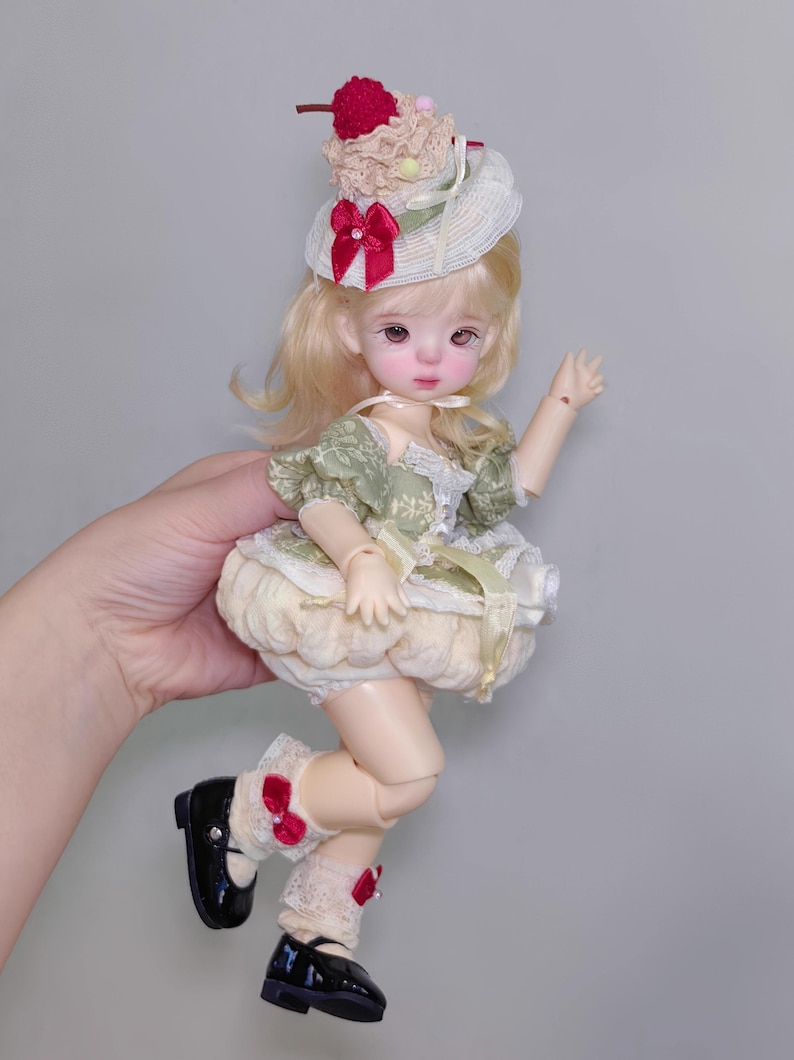 May include: A doll with blonde hair and a detailed face, wearing a green and cream-colored dress, black shoes, and a hat with a cherry on top. The doll is posed in a playful stance, with one arm raised.
