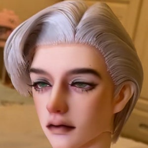 May include: A doll head with a short, silver-grey wig styled in a side part with a wave. The doll has realistic features, including green eyes and a neutral expression.