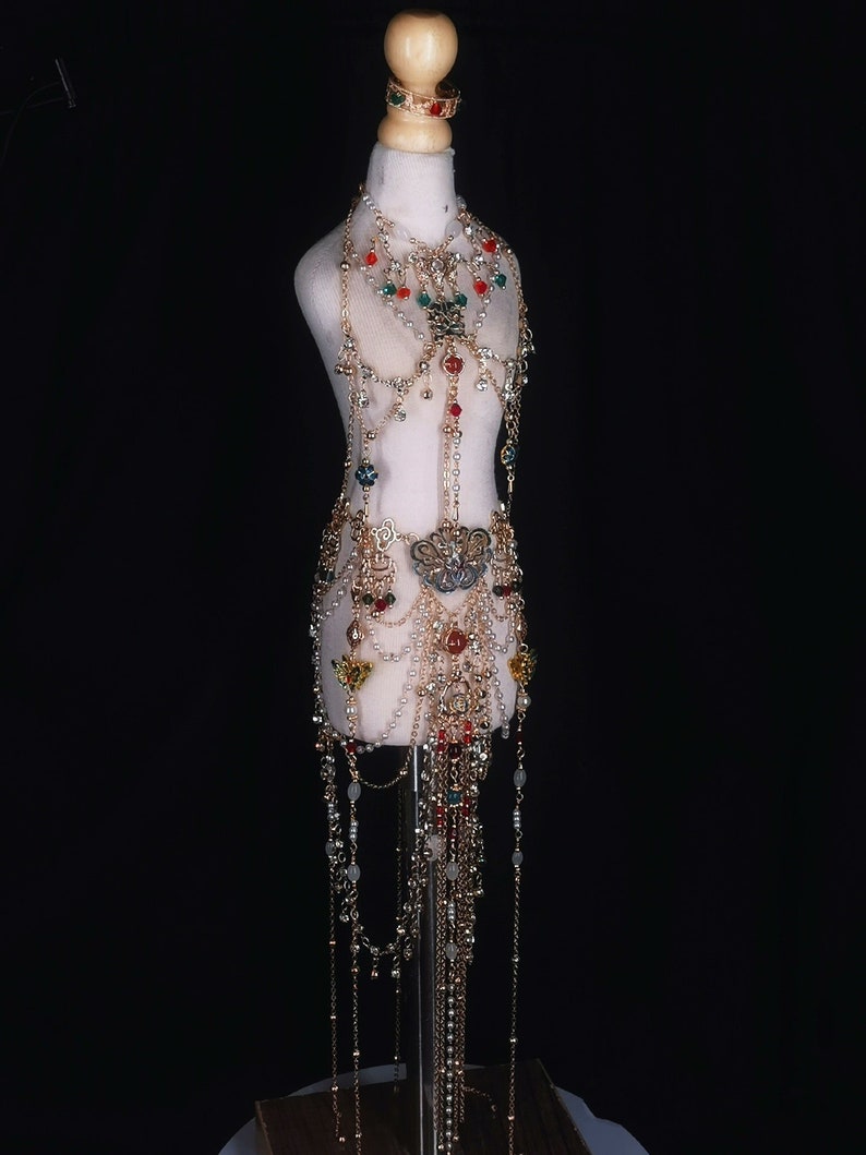 May include: A gold body chain with colorful beads and a large ornate pendant. The chain is layered with multiple strands and has a long, flowing design.