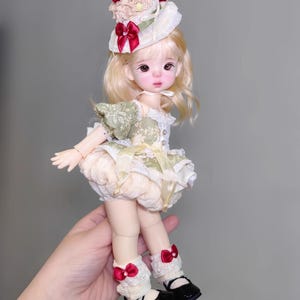 May include: A doll with blonde hair and a whimsical outfit. The doll wears a hat with a cherry on top, a green and cream dress, and black shoes. Red bows adorn the doll's socks. The doll is held in a hand.