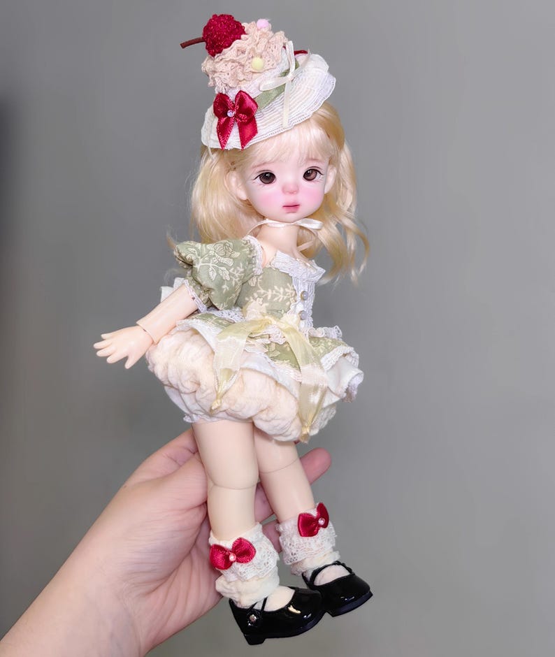 May include: A doll with blonde hair and a whimsical outfit. The doll wears a hat with a cherry on top, a green and cream dress, and black shoes. Red bows adorn the doll's socks. The doll is held in a hand.
