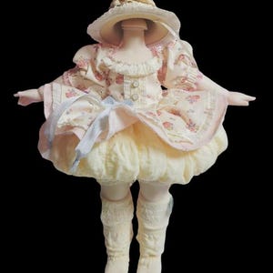 May include: A porcelain doll wearing a cream-colored dress with a floral pattern, a matching hat, and lace trim. The doll's dress has a ruffled skirt and is adorned with ribbons. The doll is wearing cream-colored socks.