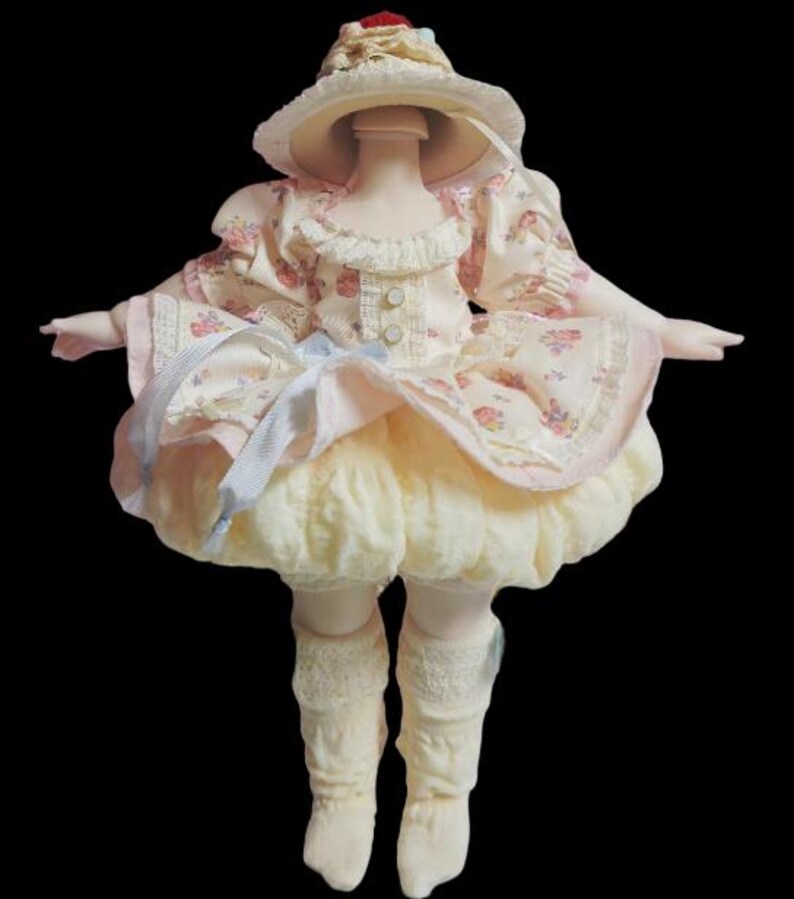 May include: A porcelain doll wearing a cream-colored dress with a floral pattern, a matching hat, and lace trim. The doll's dress has a ruffled skirt and is adorned with ribbons. The doll is wearing cream-colored socks.