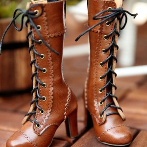 May include: A pair of brown leather boots with a lace-up design. The boots have a high heel and decorative stitching along the sides. The laces are black, and the eyelets are gold-coloured. The boots are displayed on a wooden surface.
