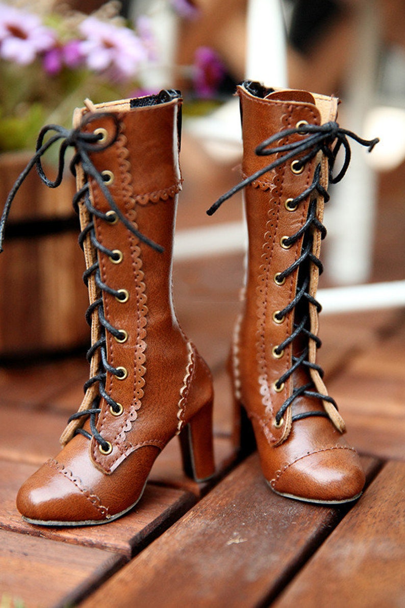 May include: A pair of brown leather boots with a lace-up design. The boots have a high heel and decorative stitching along the sides. The laces are black, and the eyelets are gold-coloured. The boots are displayed on a wooden surface.