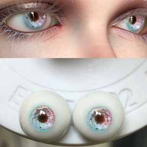 May include: Close-up of two realistic doll eyes with a blue, pink, and brown iris. The eyes are on a white surface with the number "2" visible.
