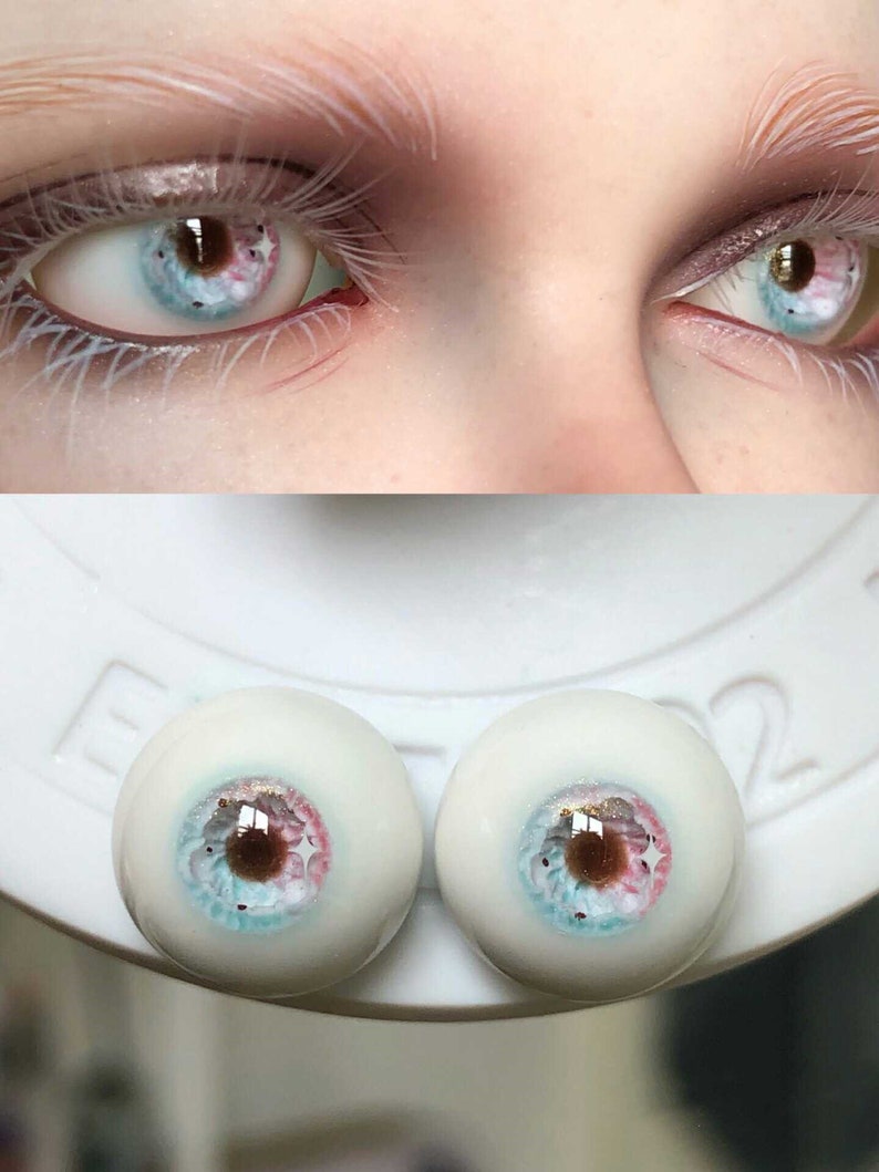 May include: Close-up of two realistic doll eyes with a blue, pink, and brown iris. The eyes are on a white surface with the number "2" visible.