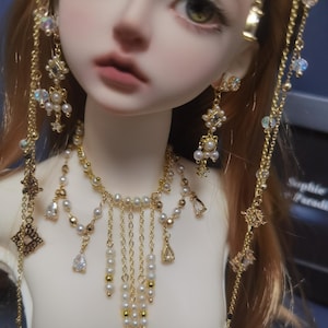 May include: A doll wearing a gold-toned necklace and matching earrings. The necklace features a multi-strand design with white pearls, gold beads, and clear teardrop-shaped crystals. The earrings are long, dangling pieces with similar pearls and crystals.