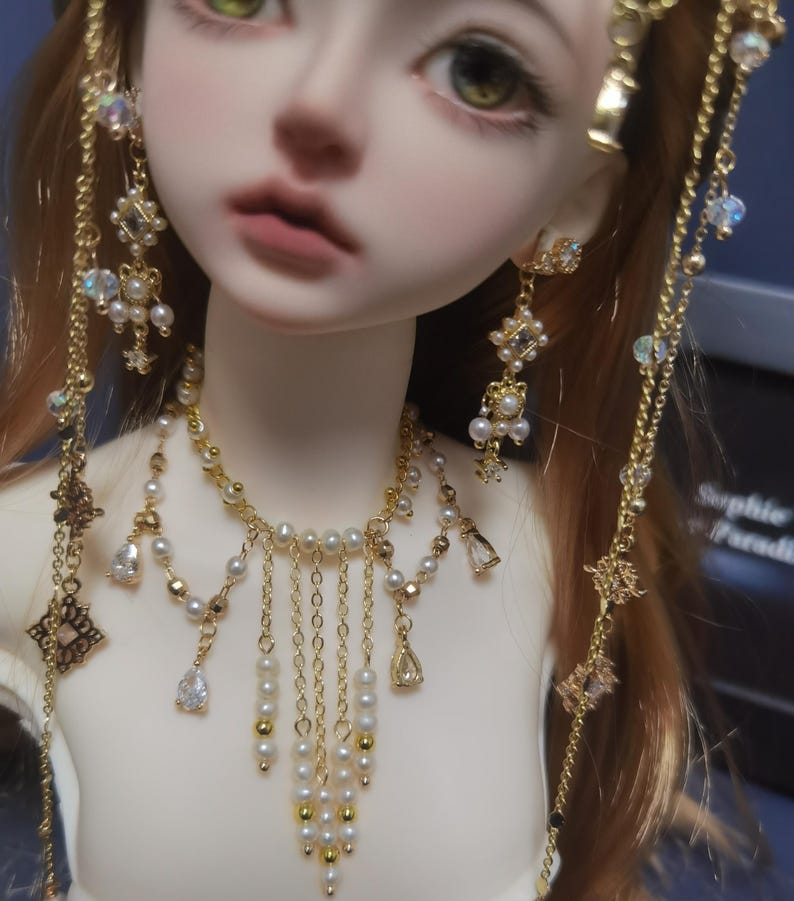 May include: A doll wearing a gold-toned necklace and matching earrings. The necklace features a multi-strand design with white pearls, gold beads, and clear teardrop-shaped crystals. The earrings are long, dangling pieces with similar pearls and crystals.