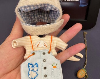 May include: A doll outfit with a white bunny hat, a yellow and white striped shirt, blue overalls with a bunny patch, and a carrot pocket. The overalls have a 'HELLO' tag on the bottom. The outfit comes with a brown bread charm on a gold chain.