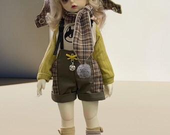 May include: A doll wearing a green hooded jacket with cat ears, a plaid scarf, a brown and white plaid overall, and white sneakers. The doll has blonde hair and blue eyes.