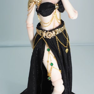 May include: A black and gold doll outfit with a gold chain belt, a gold chain necklace with green gems, and a gold chain armlet with green gems.