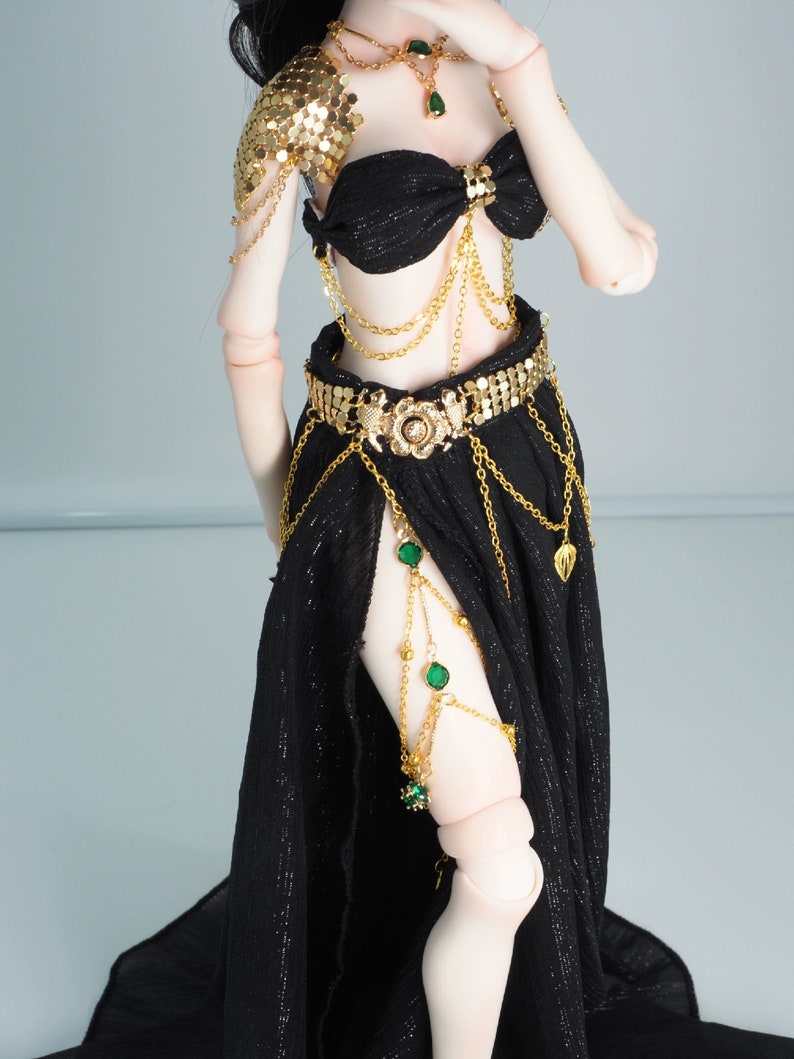 May include: A black and gold doll outfit with a gold chain belt, a gold chain necklace with green gems, and a gold chain armlet with green gems.