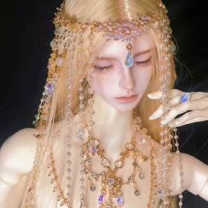 May include: A doll with long blonde hair wearing a gold and crystal crown and necklace. The doll is wearing a white dress and has a delicate face.
