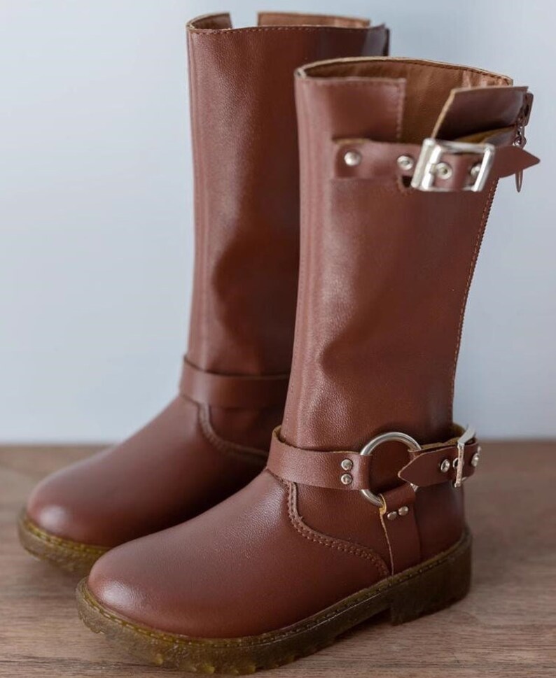 May include: A pair of brown leather knee-high boots with silver buckles and a decorative strap with a silver ring.