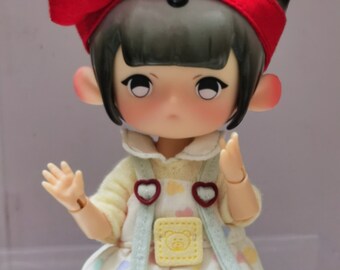 May include: A small doll with black hair and a red bow headband. The doll is wearing a light blue and white dress with a yellow patch and a bear design. The doll is holding out its hands.