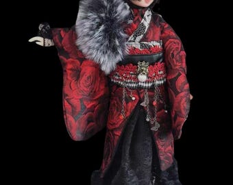 May include: A doll wearing a red kimono with a rose pattern, a black belt, and a faux fur collar. The doll has a black wig, a red and black head covering, and a decorative eye mask. The doll's outfit includes a black lace skirt and silver chain accents.
