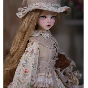 May include: A porcelain doll with long blonde hair wearing a beige dress with floral print and a white hat with floral trim.