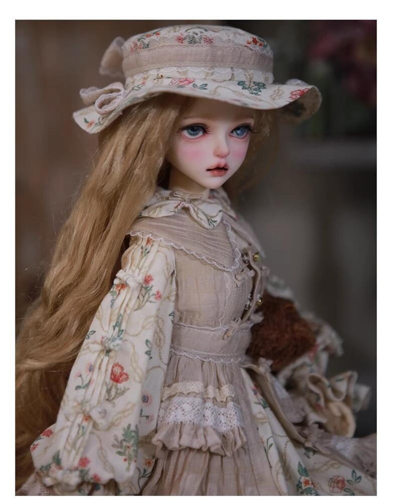 May include: A porcelain doll with long blonde hair wearing a beige dress with floral print and a white hat with floral trim.