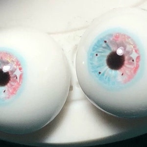 May include: Two realistic-looking glass eyeballs with blue, pink, and white painted irises and black pupils. The eyeballs are on a white surface.