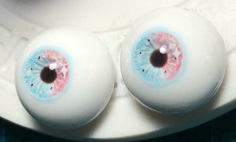 May include: Two realistic-looking glass eyeballs with blue, pink, and white painted irises and black pupils. The eyeballs are on a white surface.