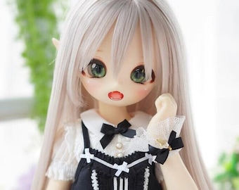Puede incluir: Una muñeca con el pelo largo y blanco que lleva un vestido blanco y negro con un cuello blanco y un lazo negro. La muñeca tiene ojos verdes y una boca rosa.